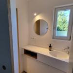 Badezimmer mit Waschbecken, runden Spiegel und Fenster mit Blick auf Bäume.