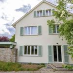 Ein zweistöckiges Haus mit hellen Wänden und grünen Fensterläden. Vor dem Haus wachsen Bäume und Sträucher.