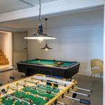 Raum mit Tischfußball und Billardtisch unter Hängelampen. Treppenhaus im Hintergrund.