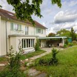 Ein zweistöckiges Haus mit Balkon, Garten und Terrasse unter einem bewölkten Himmel.