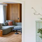 Ein Wohnzimmer mit grauem Sofa, Holzboden und Holzwand. Fenster mit Vorhängen.
