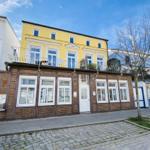 Gelbes Haus mit Balkon und rotem Backsteinbau an Straße mit Kopfsteinpflaster