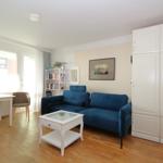 Living room with blue sofa, white furniture, and wooden floor. Windows and curtains visible.