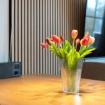 Ein Glas mit Tulpen steht auf einem Holztisch. Im Hintergrund ist ein Fernseher und eine Wandverkleidung zu sehen.
