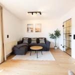 Wohnzimmer mit Sofa, Tisch und Pflanze. Holzboden und Fenster mit Vorhängen.