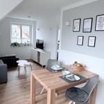 Essbereich mit Holztisch und Stühlen. Wand mit Bildern. Wohnbereich mit TV und Fenster.