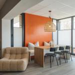 Ein Raum mit Essbereich, Sofa und orange Wand. Große Fenster und hängende Lampe.