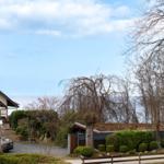 Haus mit Garten, Zaun und Bäumen unter blauem Himmel.