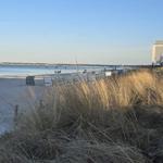 Strand mit Dünen, Steg und Hotel im Hintergrund.