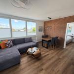Wohnzimmer mit Sofa, Esstisch und Schlafzimmer im Hintergrund. Holzboden und Backsteinwand.