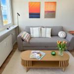 Ein Wohnzimmer mit grauem Sofa, Holztisch und Blick aufs Meer.