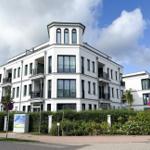 Modernes weißes Mehrfamilienhaus mit Balkonen und grüner Hecke an der Straße.
