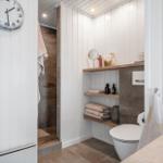 Badezimmer mit Dusche, Toilette und Holzregalen. Wanduhr und Handtücher sichtbar.