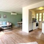 Living area with sofa, table, and kitchen. Wooden floor, green walls, windows with view of greenery.