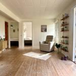 Living area with armchair, wooden floor, and view into bedrooms. Shelving and window visible.