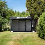 Ein weißer Schuppen mit schwarzen Rahmen und Schild Stendyssen steht im Gras zwischen Bäumen.