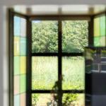 Durch ein Fenster mit bunten Glasflächen blickt man auf einen grünen Garten mit Bäumen.