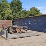 Holztische und Bänke auf einer Terrasse vor blauen Ställen mit Bäumen im Hintergrund.