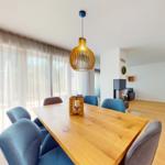 Esszimmer mit Holztisch, blauen Stühlen und Deckenleuchte. Durchgehende Fensterfront mit Gardinen.