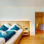 Schlafzimmer mit Bett, Holzrahmen und blauer Decke. Blick in angrenzenden Raum.