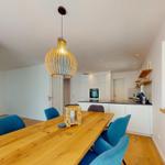 Essbereich mit Holztisch, blauen Stühlen und einer Deckenlampe. Im Hintergrund Küche und Kamin.
