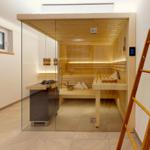 Sauna mit Holzbank, Glaswand und digitaler Steuerung. Holzstufen und Fenster sichtbar.