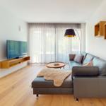 Wohnzimmer mit Sofa, Fernseher und Holzboden. Große Fenster mit Vorhängen.