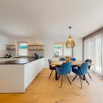 Moderne Küche mit Essbereich und großem Fenster. Holzboden und blaue Stühle.