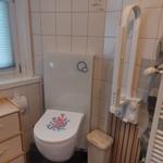 Bad mit Toilette, Duschbereich und Heizung. Fenster mit Jalousie und Blumen.