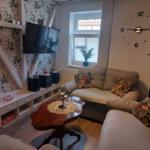 Wohnzimmer mit Sofa, Fernseher und Tisch. Wanduhr und Pflanzen sichtbar.