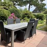 Outdoor area with covered table and six chairs. Surrounded by flowers and greenery.