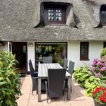 Terrace with table and chairs in front of a house with thatched roof and flower beds.