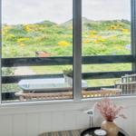 Large window with view of green hills and yellow flowers. Balcony with table and chairs.