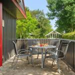 Terrace with table and chairs, red door, wooden railing, green trees in the background.