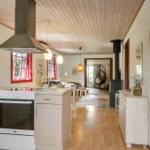 Kitchen with stove and oven, wooden ceiling, red windows, view into living area with seating.