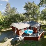 Red wooden house with terrace, blue umbrella, and garden surrounded by trees.