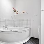 Bad mit Ecke-Badewanne, weiße Fliesenwand und kleinem Regal mit Pflanze.