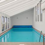 Innenpool mit blauem Wasser, weißen Wänden und Fenstern, mit Holzstühlen und Metallstufen.