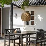 Terrasse mit Sitzgelegenheiten und Holztisch unter Glasdach. Hängelampe und Pflanzen sichtbar.