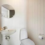 Badezimmer mit Toilette, Waschbecken und runden Spiegel an weißen Holzwänden.