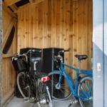 Zwei Fahrräder stehen in einem Holzstall. Ein schwarzes mit Korb, ein blaues.