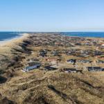 Aerielansicht von Strand, Dünen und Häusern mit Windkraftanlagen im Hintergrund.