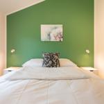 Schlafzimmer mit grüner Wand, Bett mit weißer Bettwäsche und Kissen, zwei Nachttische mit Lampen.