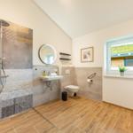 Badezimmer mit Dusche, Waschbecken und WC. Holzoptik Boden, Fenster mit Blick.