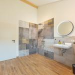 Bad mit Dusche, Waschbecken und Toilette. Fliesenwand und Holzdielenboden.