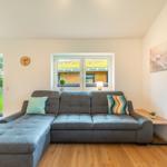Ein Wohnzimmer mit grauem Sofa, Holzboden und Fenster zum Garten.