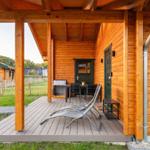 Terrasse mit Liegestühlen und Tisch unter Holzüberdachung. Hintergrund: Grasfläche und weitere Holzhäuser.