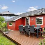 Red wooden house with terrace, table, chairs, and umbrella. Greenery and flowers surround the house.