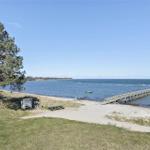 Strand mit Sand und Steinen, ruhiges Wasser und Wald im Hintergrund.