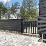Black wooden cabin with terrace, table, and chairs. Surrounded by trees and grass.